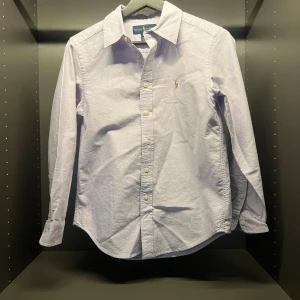 Skjorta från Ralph Lauren - Snygg ljusblå/lila skjorta från Ralph Lauren med klassisk krage och broderad logotyp på bröstet. Skjortan har långa ärmar och knappar framtill. Perfekt för en stilren look.