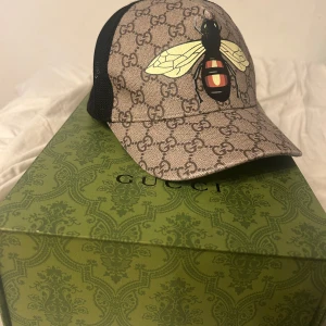 Gucci keps med bi-motiv - Snygg keps från Gucci med ett coolt bi-motiv på framsidan. Kepsen har ett klassiskt GG-mönster i beige och svart mesh på sidorna. Perfekt för att ge din outfit en lyxig touch.