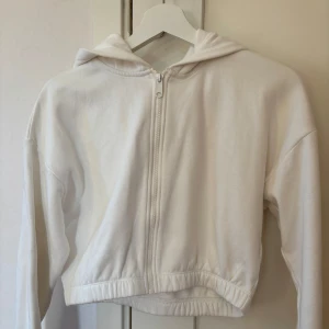 Vit croppad hoodie från Lindex  - Säljer en stilren vit croppad hoodie med dragkedja. Perfekt för en avslappnad look. Den har långa ärmar och en bekväm huva. Passar bra till jeans eller leggings.
