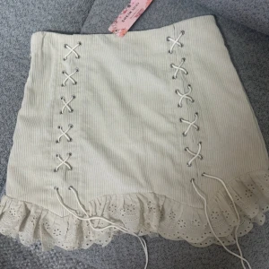 Begie  ribbad kjol med snörning - Söt vit ribbad kortkjol med snörning på sidorna och volangdetalj i nederkant. Perfekt för en somrig look. 💕