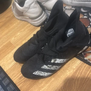 Svarta Adidas Predator fotbollsskor - Säljer ett par svarta Adidas Predator fotbollsskor med vita detaljer. Skorna har snörning och är designade för optimal kontroll på planen. Perfekta för fotbollsspelare som vill ha både stil och funktion.