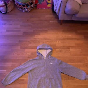 Grå hoodie från Nike - Säljer en grå hoodie från Nike med dragkedja och huva. Den har långa ärmar och en klassisk design med Nike-logga på bröstet. Perfekt för en avslappnad stil.