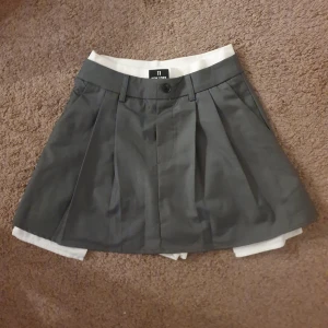 Grå kortkjol med inbyggda shorts - Snygg grå kortkjol med inbyggda vita shorts och ett par fickor. Kjolen har en knapp och dragkedja framtill samt veckade detaljer för en stilren look. Perfekt för en aktiv dag eller en avslappnad stil.