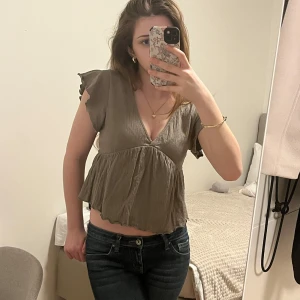 Beige blus med volangdetaljer - Säljer en söt beige blus med volangdetaljer och v-ringning. Blusen har korta ärmar och en luftig passform, perfekt för varmare dagar. Den är tillverkad i ett lätt och bekvämt material.