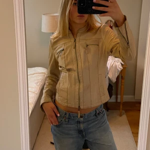Beige skinnjacka med dragkedja - Snygg beige skinnjacka med dragkedja framtill och flera praktiska fickor med dragkedjor. Jackan har en stilren design och är perfekt för en cool och avslappnad look. Passar bra till jeans och andra casual outfits.