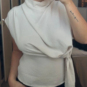 Vit omlottopp - Säljer en stilren vit omlottopp med knytning i sidan. Toppen har en unik design med en ärm och en hög krage, vilket ger en elegant look. Perfekt för att lyfta vilken outfit som helst.