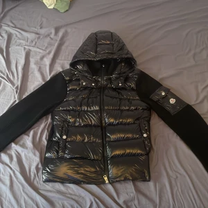 Svart cardigan från Moncler - Säljer en snygg svart cardigan från Moncler med huva och dragkedja. Jackan har långa ärmar och en glansig finish. Perfekt för kyligare dagar med stil. Det e storlek L men passar M för dom e ganska små i storlekar 
