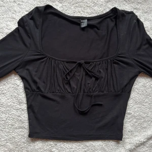 Svart långärmad topp från Shein - Säljer en stilren svart långärmad topp från Shein. Toppen har en söt knytning framtill och en fyrkantig urringning som ger en elegant touch. Perfekt för att matcha med både jeans och kjol. Passar bra för både vardag och festliga tillfällen.