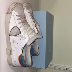 Vita sneakers från Philippe Model - Snygga vita sneakers från Philippe Model med grå detaljer och en blå och orange häl. Skorna har snörning och en röd sula. Perfekta för en stilren look. Skorna har en menad ”beat up” look som bidrar till en riktig fet outfit. Sko storleken är it 41 och innermått 26.5 cm. Priset är ej hugget i sten kom gärna med prisförslag.