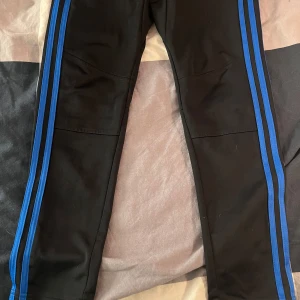 Svarta träningsbyxor från Adidas - Snygga svarta träningsbyxor från Adidas med blå ränder längs sidorna. Byxorna har en elastisk midja med snörning och är tillverkade i Climalite-material för optimal komfort under träning. Perfekta för sport och aktivitet.