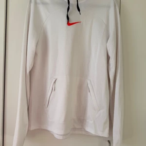 Vit hoodie från Nike - Säljer en vit hoodie från Nike med röd logga på bröstet. Tröjan har en huva med svarta dragsnören och en praktisk magficka. Materialet är Dri-FIT vilket gör den perfekt för träning.