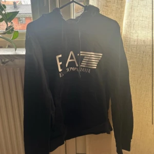 Svart hoodie från Emporio Armani - Säljer en svart hoodie från Emporio Armani med deras logga tryckt på framsidan. Tröjan har en klassisk design med huva och dragsko. Perfekt för en stilren och bekväm look.