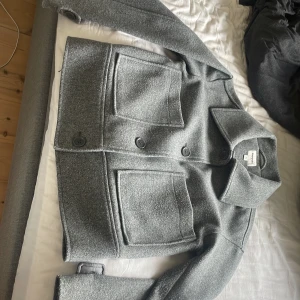 Grå kappa från Kappahl - Säljer en stilren grå kappa från Kappahl i storlek XS. Jackan har en klassisk design med stora knappar och två framfickor. Perfekt för kyligare dagar.