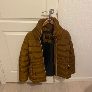 Brun dunjacka från Lindex - Säljer en snygg brun dunjacka från Lindex. Jackan har en dragkedja framtill och praktiska fickor med dragkedjor. Perfekt för kyligare dagar med sin vadderade design och höga krage.