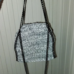 Silvergrå virkad väska med kedjedetaljer - Snygg silvergrå väska med en unik textur och svarta kedjedetaljer. Perfekt för att ge din outfit en edgy touch. Väskan har en stilren design och är lätt att matcha med olika stilar.