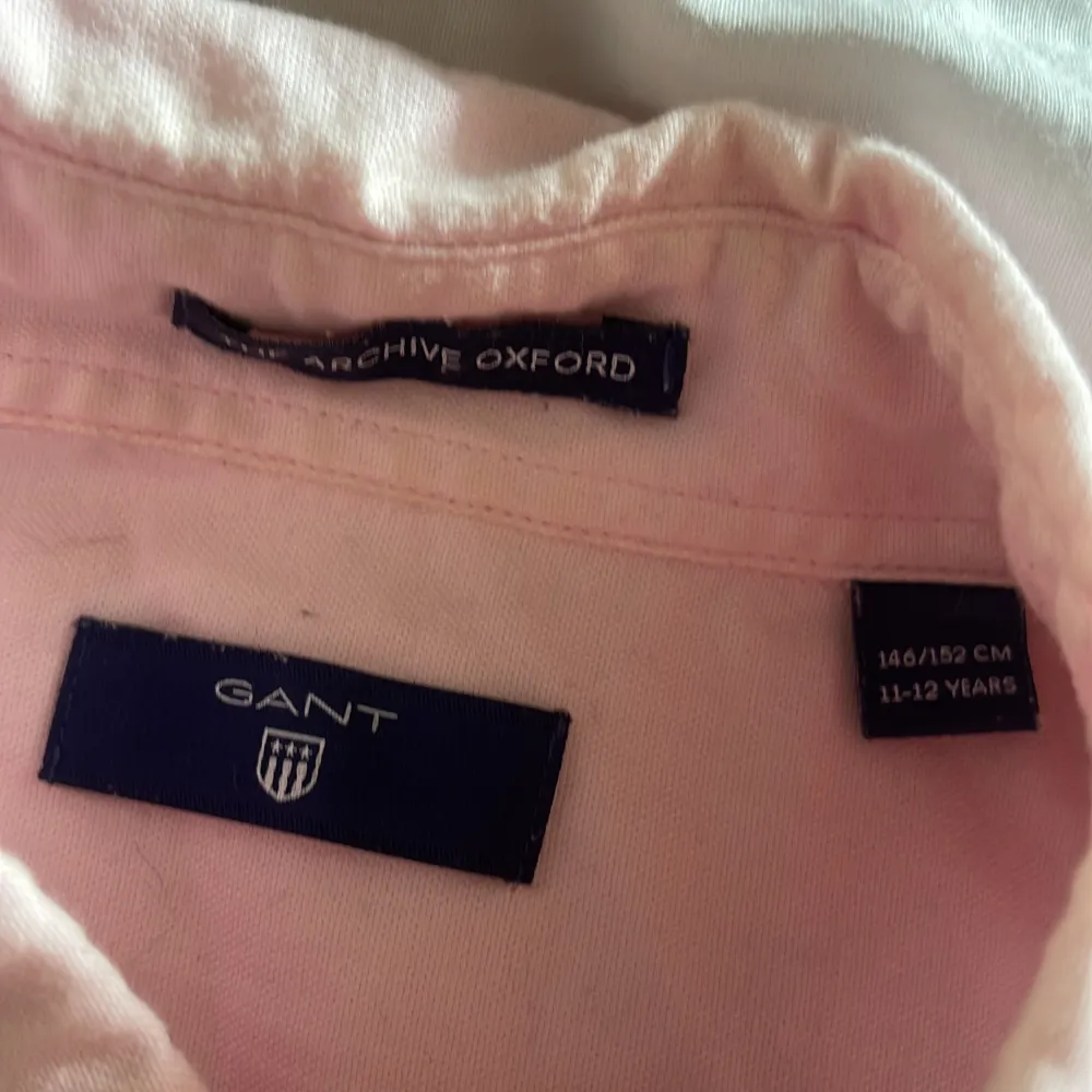 Säljer en stilren rosa skjorta från GANT med broderad logga på bröstfickan. Skjortan har långa ärmar och knappar framtill. Perfekt för en klassisk och avslappnad look.. Paidat.