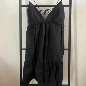 Svart klänning med smala axelband - Snygg svart klänning med smala axelband och v-ringning. Klänningen har en volangdetalj nedtill och knytning i ryggen. Perfekt för en stilren look.