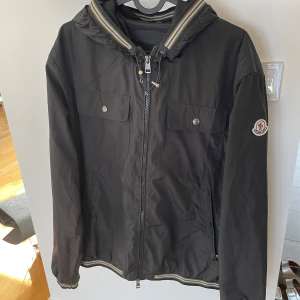 Svart vindjacka från Moncler - Säljer en stilren svart vindjacka från Moncler med huva och dragkedja. Jackan har två bröstfickor med knappar och Moncler-logga på ärmen. Perfekt för blåsiga dagar.  Säljer pga att den börjar bli för liten. 