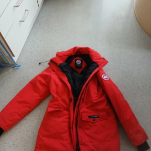 Röd dunjacka från Canada Goose - Säljer en röd dunjacka från Canada Goose med lång ärm och dragkedja. Jackan har en klassisk design med en logotyp på ärmen och flera praktiska fickor. Perfekt för kalla vinterdagar.