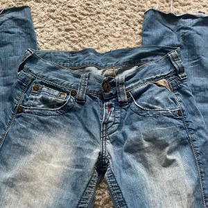 Snygga replay jeans - Säljer ett par blå jeans från Replay med snyggt slitna detaljer och en broderad orange patch med lila motiv bak på linningen. Jeansen har raka/lite bootcut klassiska fem fickor och kontrastsömmar. Perfekt för dig som gillar street och vill ha något unikt i garderoben. Storlek - 25/32 midjemåttet - 36,5 cm innerbenslängd - 81 cm