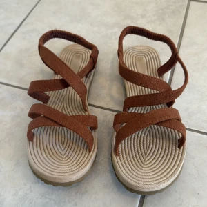 Bruna sandaler med korsade remmar - Säljer ett par snygga sandaler i storlek 39 med korsade bruna remmar i mockaliknande material och beige sula med mönstrad struktur. Remmarna går runt foten och ger en cool och avslappnad vibe, perfekt för varma dagar. Sulan är platt och bekväm för långa promenader. Oanvända då jag råkade beställa två par av samma sko.