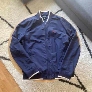 Ralph Lauren jacka - Tjena👋, säljer nu denna snygga mörkblåa jacka från Polo Ralph Lauren med klassisk röd broderad logga på bröstet🪡. Jackan har vit-randiga detaljer på krage och muddar, dragkedja framtill och två sidofickor🧥 Perfekt för dig som vill ha en stilren och sportig look🤩  Jackan säljs då den är för liten och inte används🙌 Den är barnstorlek XL men sitter som vuxenstorlek XS📐 Diskutera pris eller få mått, kom privat📩