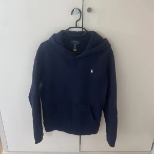 Marinblå hoodie från Polo Ralph Lauren - Snygg marinblå hoodie från Polo Ralph Lauren med klassisk vit logga på bröstet. Tröjan har huva, känguruficka och långa ärmar. Tillverkad i mjuk bomull som är skön att ha på sig. Perfekt för chill dagar och enkel att matcha med jeans eller joggers.