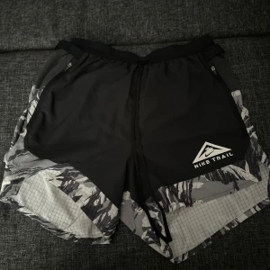 Nike Trail Dri-Fit shorts S - Nike Trail Dri-Fit shorts i svart och grått med coolt camouflage-mönster. Lätta och ventilerande med snörning i midjan och dragkedjefickor. Perfekta för löpning och träning, tillverkade i syntetmaterial som håller dig torr och bekväm.