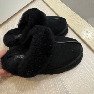 Svarta UGG boots med päls - Mysiga svarta boots från UGG med fluffig päls runt öppningen och grov platt sula. Skorna har rund tå och är tillverkade i mocka med mjukt pälsfoder som håller fötterna varma. Perfekta för kalla dagar och har en klassisk UGG-logga på sidan.