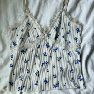 Blommig spetskantad linnetopp - Supersöt linnetopp med tunna axelband och spetskant vid halsringningen. Toppen är vit med blå blommigt mönster och har diskreta vertikala ränder. Liten rosett framtill och ribbad struktur ger extra detaljer. Perfekt för en chill sommardag.