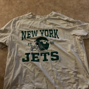Vit H&M T-shirt  - Vit t-shirt från H&M tryck i grönt och svart framtill, inklusive hjälmmotiv och årtalet 1960. Klassisk rund hals och korta ärmar. Perfekt för dig som gillar amerikansk fotboll och vill ha en sportig vibe.
