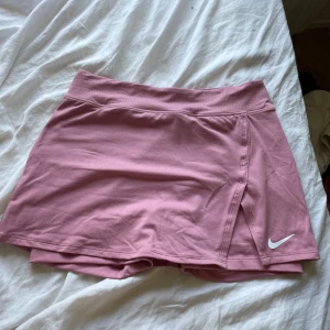 Rosa sportkjol från Nike - Säljer en snygg rosa sportkjol från Nike med inbyggda shorts under. Kjolen har en bred resår i midjan och en vit Nike-logga på framsidan. Perfekt för tennis, padel eller gymmet. Materialet är mjukt och stretchigt för maximal rörelsefrihet.