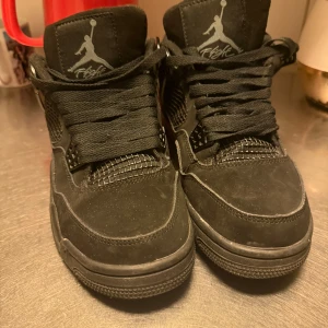 Svarta Nike Jordan 4 sneakers - Säljer ett par svarta Nike Jordan 4 sneakers med klassisk Jumpman-logga på plösen och hälen. Skorna har snörning, meshdetaljer på sidorna och platt sula. Materialet är mocka och syntet, och designen är riktigt clean och sportig. Perfekt för dig som gillar streetwear och sneakers med attityd.