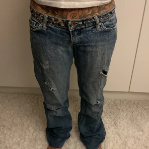 Levis 501 blå jeans med slitningar - Säljer ett par klassiska  sixsis jeans i blå tvätt med coola slitningar och hål på benen. Modellen har rak passform och låg midja, samt fem fickor och knappgylf. Perfekt för dig som gillar en avslappnad och edgy stil. Midjemått: 37xm