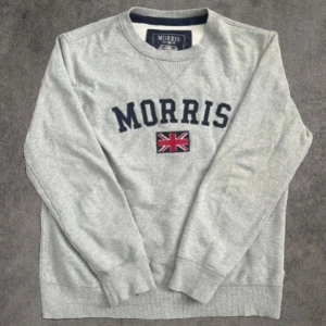 Grå sweatshirt från Morris med tryck - Snygg grå sweatshirt från Morris i storlek medium. Tröjan har ett stort Morris-tryck och en broderad brittisk flagga på bröstet. Klassisk rund hals och ribbade muddar vid ärmar och nederkant. Perfekt för dig som gillar stilren design med en brittisk touch.