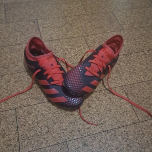 Adidas Predator fotbollsskor röd/svart - Säljer ett par Adidas Predator fotbollsskor i rött och svart med coolt rutigt mönster på ovansidan. Skorna har snörning och en låg profil med rund tå. Yttermaterialet är syntet och sulan har dobbar för gräsplan. Perfekt för dig som vill sticka ut på planen!