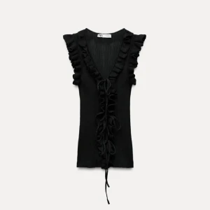 Svart ribbad topp med volang från Zara - Svart ribbad topp från Zara med djup v-ringning, volangdetaljer längs axlar och fram, samt snörning framtill. Ärmlös design som ger en edgy och trendig vibe. Perfekt att styla med jeans eller kjol för en cool look.