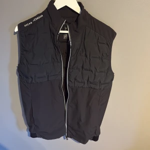 Svart dunväst från Oscar Jacobson - Svart dunväst från Oscar Jacobson med quiltad ovandel och stilren ståkrage. Västen har en vit dragkedja framtill och diskret logga på axeln och nacken. Perfekt att slänga över en hoodie eller tröja för extra värme. Snygg och enkel design som funkar till många stilar.