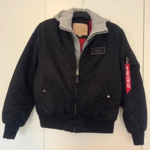Svart bomberjacka Alpha Industries M - Säljer en svart bomberjacka från Alpha Industries i storlek M. Jackan har en avtagbar grå huva med broderad logga, röd insida och klassisk patch på bröstet. Flera fickor, dragkedja framtill och rött Alpha band på ärmen. Perfekt för dig som vill ha streetstyle med edge.
