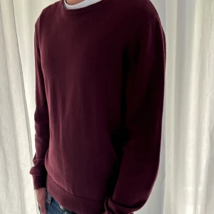 Vinröd stickad tröja med rund hals - Snygg vinröd stickad tröja med rund hals och långa ärmar. Tröjan har en enkel och clean design utan mönster, med ribbade muddar vid ärmslut och nederkant. Perfekt att matcha med jeans eller över en t-shirt för en chill look.
