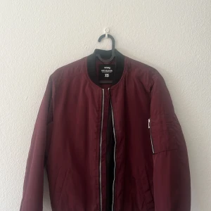 Vinröd bomberjacka från Sinsay - Säljer en vinröd bomberjacka från Sinsay med svarta ribbade muddar vid krage, ärmslut och nederkant. Jackan har dragkedja framtill och en ficka med dragkedja på ena ärmen. Perfekt för dig som gillar stilren och enkel design.