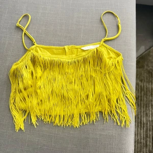 Gul croppad linnetopp med fransar - Säljer en riktigt cool gul croppad linnetopp med tunna axelband och massor av långa fransar framtill. Perfekt för dig som vill sticka ut och älskar färg! Toppen har en snygg och lekfull vibe, passar till både jeans och kjol. Fråga gärna om storlek!