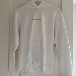 Vit hoodie med texttryck - Vit hoodie med huva och magficka, stilren design och svart texttryck 'positivité est priorité' på bröstet. Mjuk och skön, perfekt för chill dagar. Långärmad och i klassisk passform. Materialet känns som bomull eller polyester.