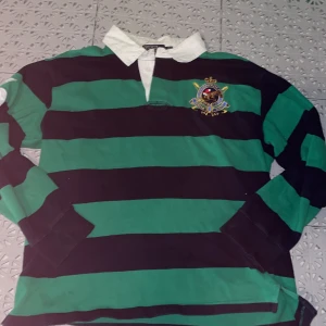 Långärmad piké från Ralph lauren - Säljer en grön och marinblå randig pikétröja från Polo Ralph Lauren i storlek L. Tröjan har vit krage, broderat emblem på bröstet och stora siffror på ryggen och ärmen. Tillverkad i 100% bomull och har långa ärmar. Perfekt för dig som gillar klassisk preppy stil. Tröjan är liten i storleken, passar mig som är S