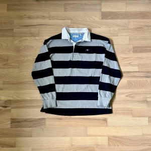 GANT piketeöja - Snygg pikétröja från GANT med breda marinblå och grå ränder, klassisk vit krage och broderad logga på bröstet. Tröjan har långa ärmar och är gjord i mjuk bomull, perfekt för en chill och sportig stil.