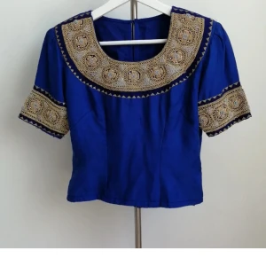 Blå topp med guld broderi - Unik blå blus / topp med korta ärmar och detaljerat guldstål broderi runt halsringningen och på ärmarna. Blusen har en rak passform och stängs med dragkedja bak. Passar storlek S-M.