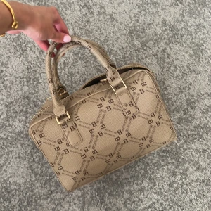 Beige handväska - Säljer en beige handväska från Bruno Banani med monogrammönster och guldfärgade detaljer. Väskan har både handtag och justerbar axelrem, samt dragkedja upptill. Insidan har fack för småsaker. Perfekt storlek för det viktigaste. Förlåt för dåliga bilder min kamera e bajs hehe.