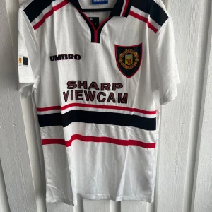 Manchester United Beckham 7 Umbro tröja - Säljer en klassisk Manchester United fotbollströja från Umbro med Beckham och nummer 7 på ryggen. Tröjan är vit med svarta och röda detaljer, klubbmärke på bröstet och SHARP VIEWCAM-tryck. Materialet är polyester och har en snygg krage med röd och svart kant.