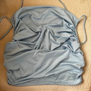 Ljusblå bandeau tubtopp med snörning - Supersnygg ljusblå bandeau tubtopp med smala axelband och cool snörning på sidorna. Toppen har en croppad passform och är gjord i mjukt, stretchigt syntetmaterial som sitter skönt mot kroppen. Perfekt för dig som vill ha en enkel men trendig look.