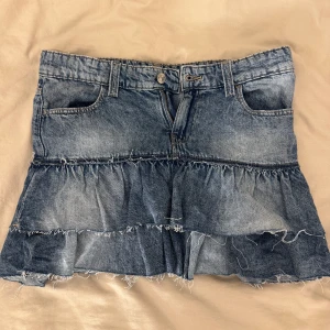 Blå jeanskjol med volanger - Säljer en kort jeanskjol i blå denim med två lager volanger och fransiga kanter. Kjolen har klassiska fickor fram men inga bak. Som helt ny. Storleken är 164